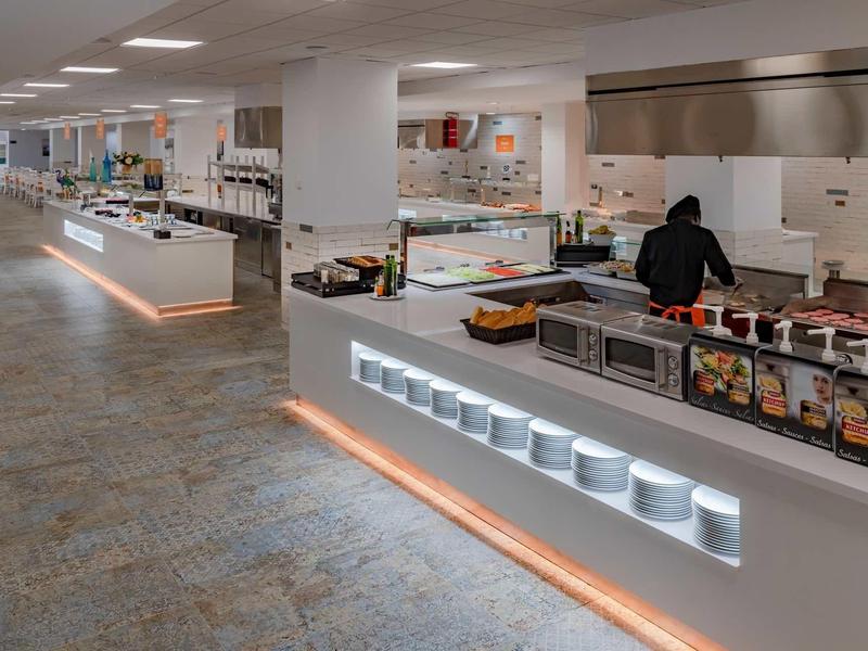 Cocina moderna de hotel con mostradores blancos, estación de cocina y amplia iluminación.