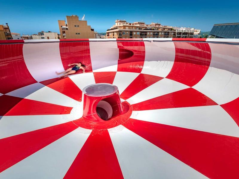 Piscina de tobogán de agua a rayas rojas y blancas bajo un cielo claro en un hotel.