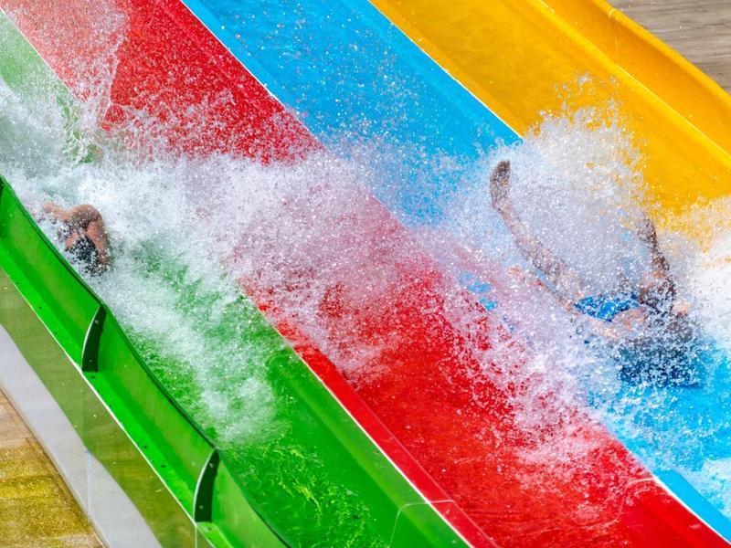 Dos personas descienden por toboganes de agua de colores amarillo, azul, rojo y verde en un parque acuático.
