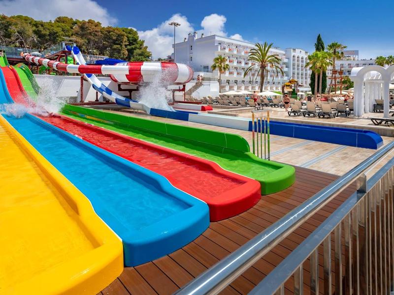 Toboganes coloridos en un parque acuático con áreas de descanso y palmeras al fondo.