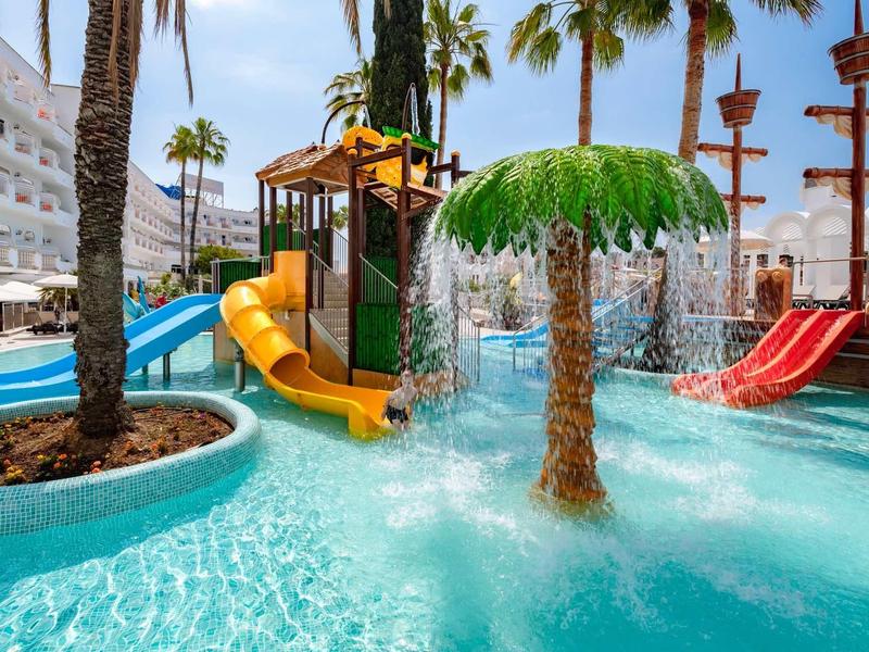 Parque acuático colorido con toboganes y palmeras en la piscina de un hotel bajo el sol.