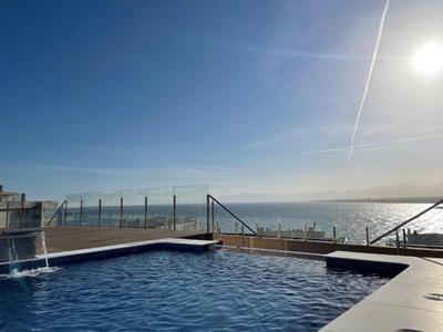 Ein rechteckiger Pool mit Wasserfall, dahinter Meer und klarer blauer Himmel mit Sonne.