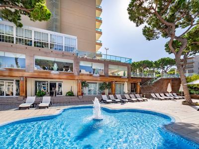 Modern hotel pool with sun loungers and fountain in a sunny garden area.