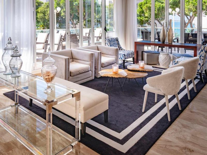 Moderní hotelový salon s pohodlnými křesly a velkým kobercem u oken od podlahy ke stropu.