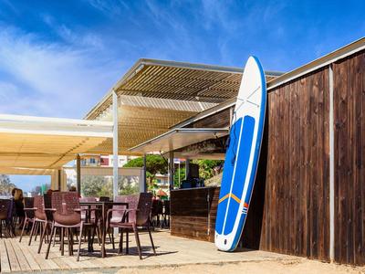 Area esterna di un bar con sedie e una tavola da surf appoggiata a una parete di legno.