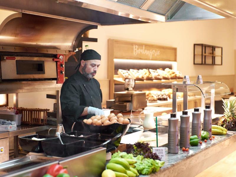 Fornaio serve panini freschi dietro un banco con frutta e salse.