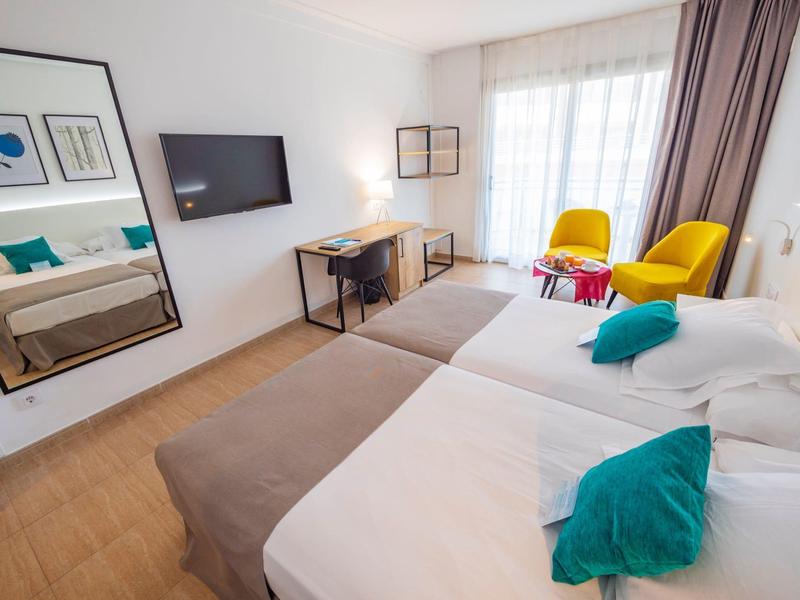 Camera d'hotel luminosa con due letti singoli, grande specchio, TV e scrivania davanti a una finestra con tende.