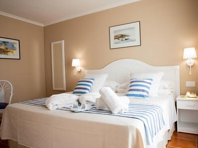 Světelný hotelový pokoj s manželskou postelí, bílým povlečením a modrobílými pruhovanými polštáři, dvě noční stolky.