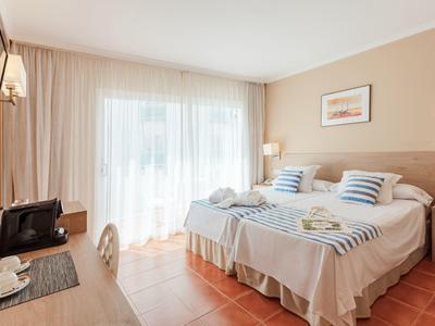 Světle zařízený hotelový pokoj se dvěma postelemi, psacím stolem a velkým oknem se závěsy.