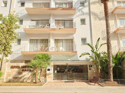 Fasáda hotelu s balkony, palmami a krytým vchodem u chodníku.