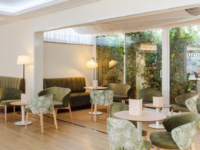 Světý hotelový pokoj se zelenými křesly, pohovkami a velkými okny s výhledem na zeleň.