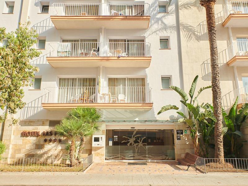 Fasáda hotelu s balkony, palmami a krytým vchodem u chodníku.