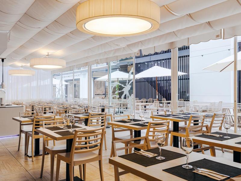 Moderní světlá restaurace s dřevěnými židlemi a prostřenými stoly pod velkým svítidlem.