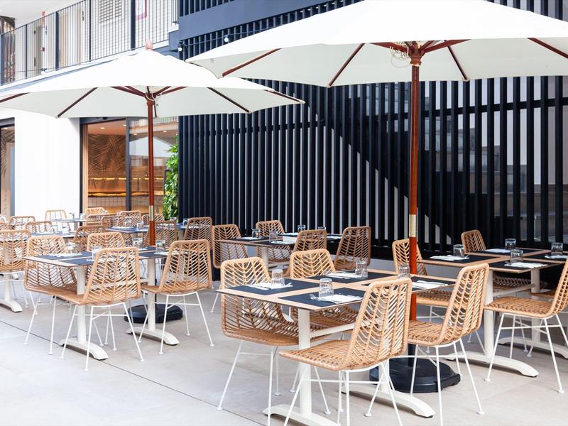 Moderní venkovní prostor kavárny se dřevěnými židlemi, stoly a velkými slunečníky.