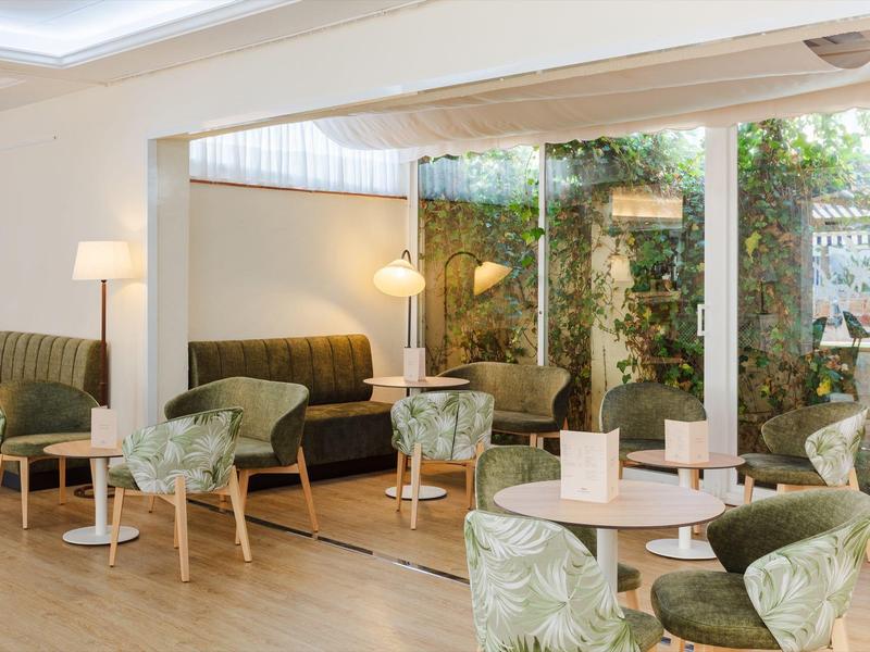 Světý hotelový pokoj se zelenými křesly, pohovkami a velkými okny s výhledem na zeleň.