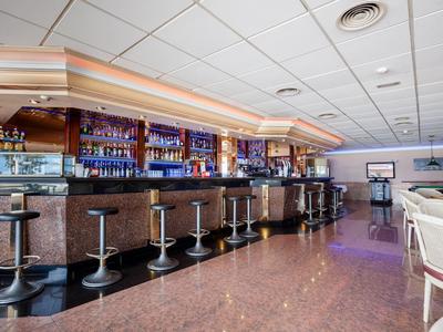 Moderne Bar mit schwarzen Barhockern, gläsernem Tresen und Spiegelwand im Deckenlicht.