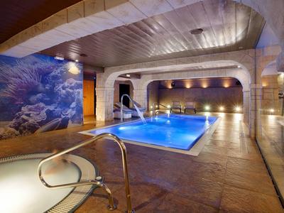 Moderner Innenpool mit blau beleuchtetem Wasser, Mosaikwand und Holzdecke in warmem Licht.