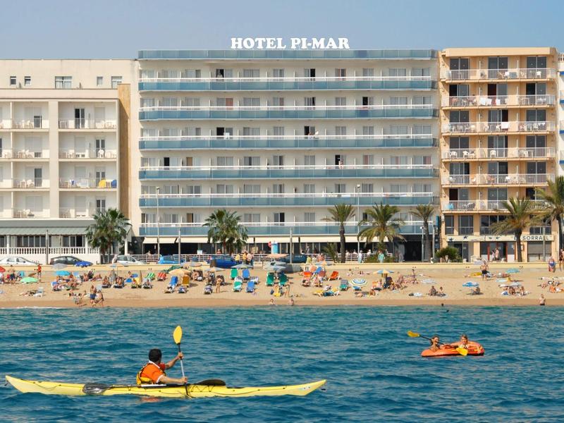 Mehrstöckiges Hotel am Strand mit Sonnenbadenden, Palmen und Kajakfahrern im blauen Meer.