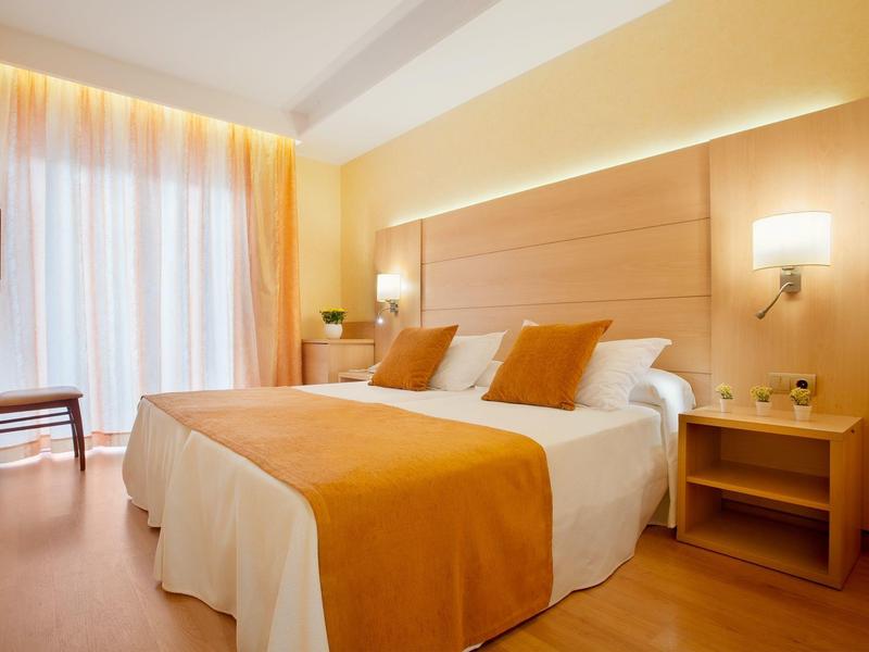 Helles Hotelzimmer mit Doppelbett, orangenem Bettüberwurf und Holzmöbeln, Fenster mit Vorhängen.