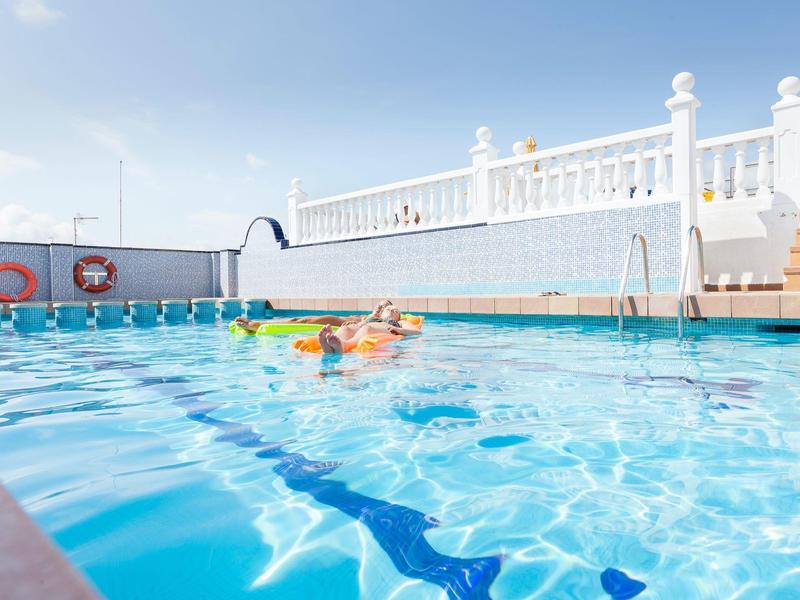 Klares Schwimmbecken mit blauer Markierung, Person schwimmt, weiße Umzäunung und blaue Himmel.