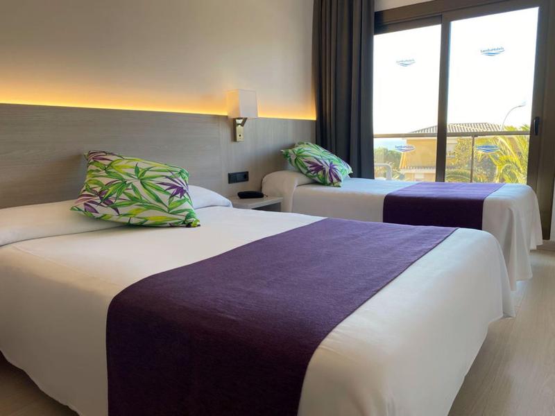 Two single beds with white linens, purple runners, and floral pillows in a bright hotel room.