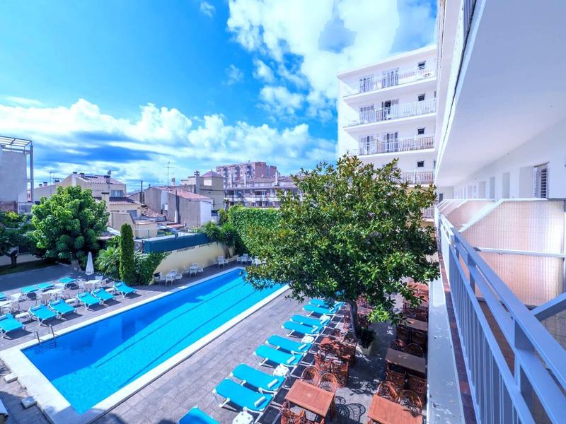 Grande piscina esterna con lettini accanto a un edificio alberghiero multi-piani sotto un cielo azzurro.