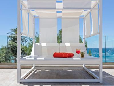 Moderner Lounge-Bettbereich mit weißem Gestell, rotem Handtuch und Blick auf Meer und Palmen.