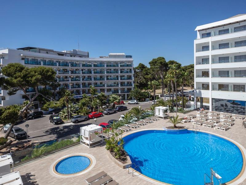 Complexe hôtelier avec grandes piscines, terrasse ensoleillée et bâtiments environnants sous un ciel bleu.