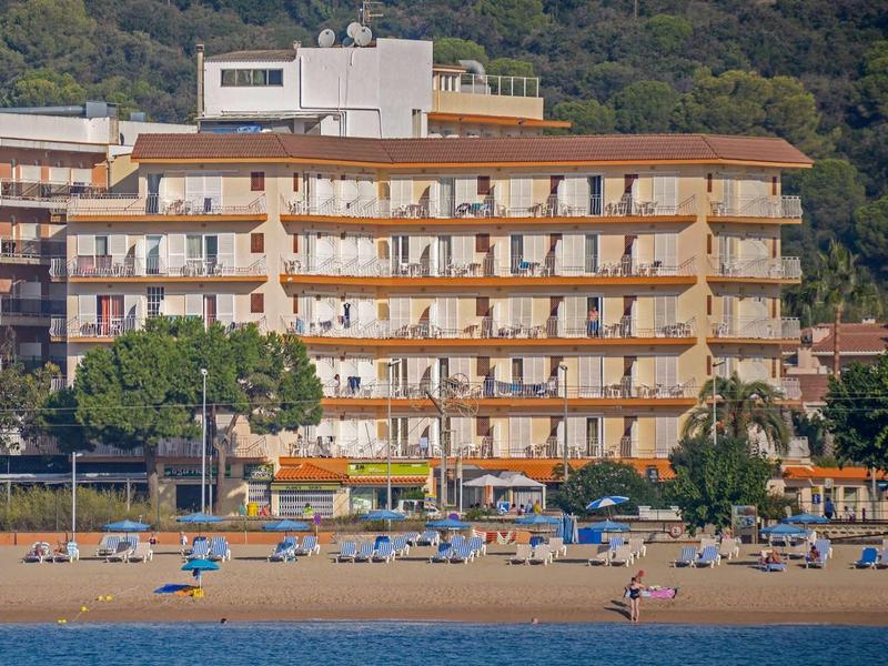 Wielopiętrowy hotel z balkonami skierowanymi na plażę, leżaki i parasole na piasku.