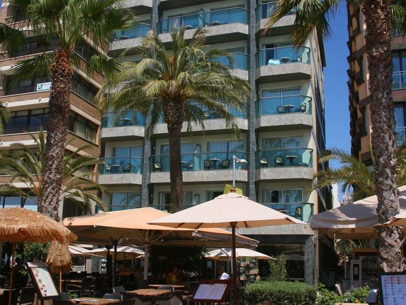 Multi-story hotel with balconies, palm trees, and outdoor seating with tables and umbrellas.