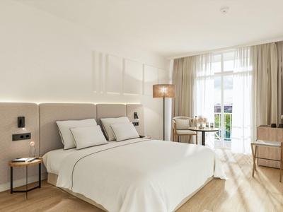 Světelný pokoj s velkou postelí, kulatým stolkem, stojací lampou, psacím stolem a balkonem.