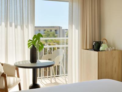 Světle zařízený pokoj s balkonem, kulatý stůl s rostlinou a světlé závěsy.