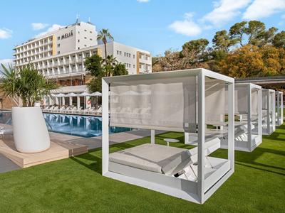 Bílé venkovní postele u bazénu a hotelu pod jasně modrou oblohou.