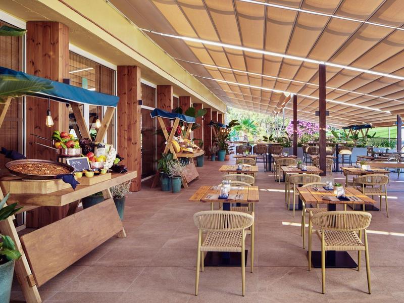 Venkovní kavárenská terasa s stolky a židlemi pod béžovou markýzou.