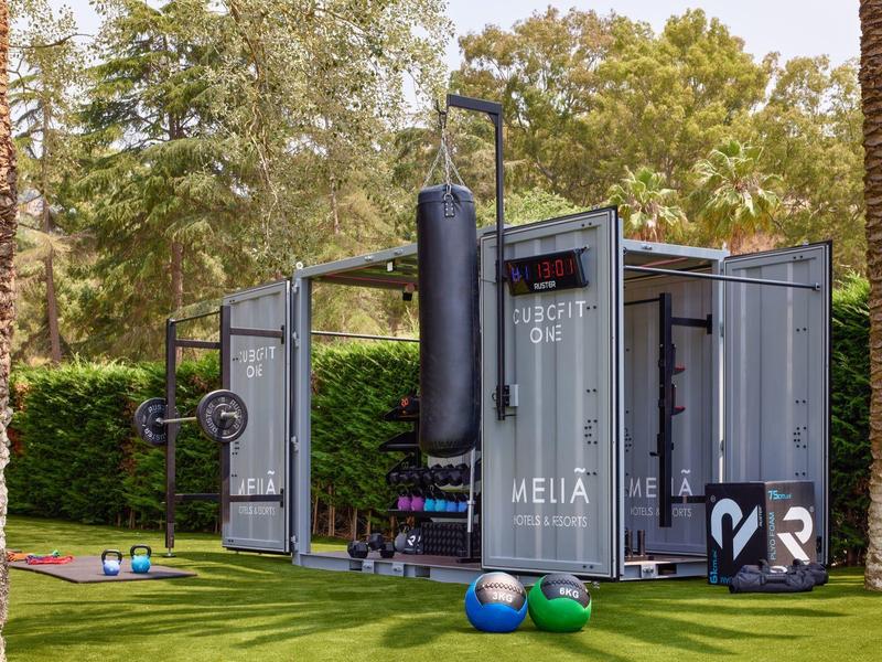 Kontejner přeměněný na venkovní fitness prostor s vybavením na zeleném trávníku.