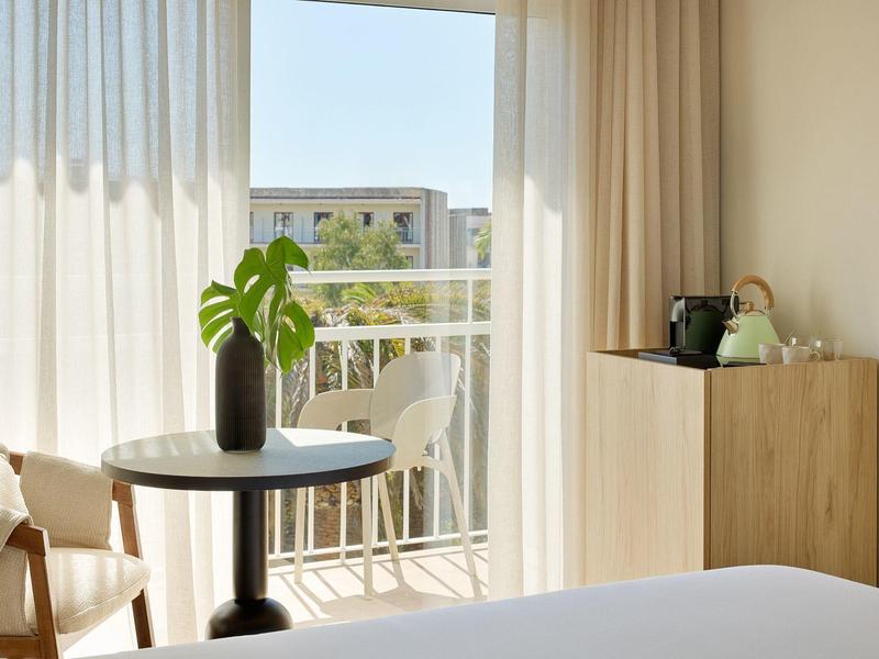Světle zařízený pokoj s balkonem, kulatý stůl s rostlinou a světlé závěsy.