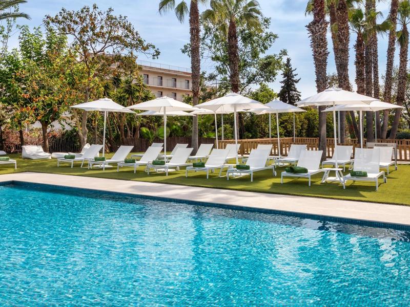 Světle modrý bazén obklopený bílými lehátky a slunečníky pod palmami.