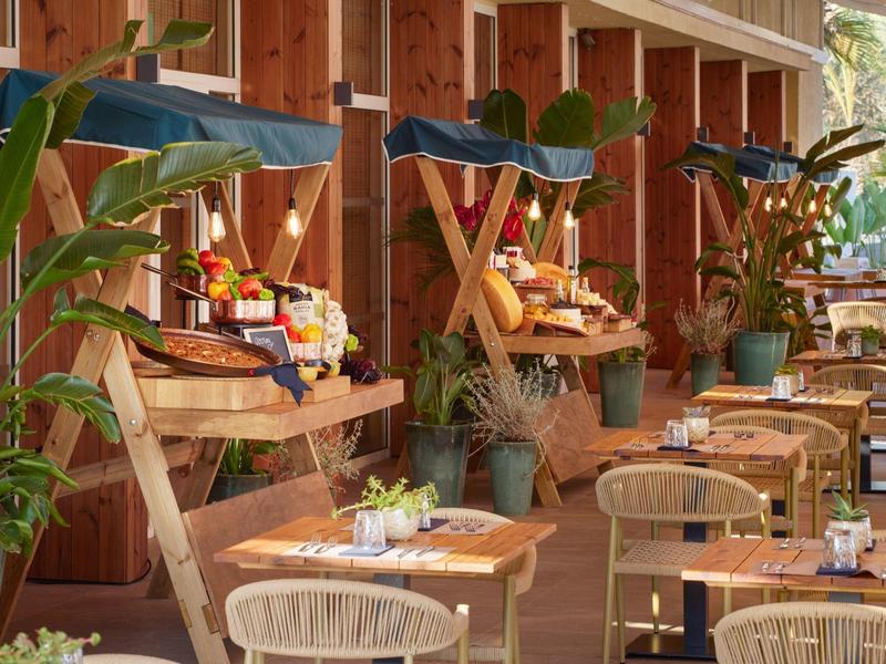 Dřevěná terasa s stolky, židlemi a velkými pohodlnými křesly, obklopená zelenými rostlinami.