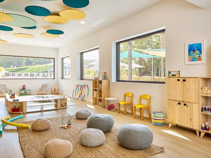 Světelná herna s polštáři, velkými okny a světlým dřevěným nábytkem.