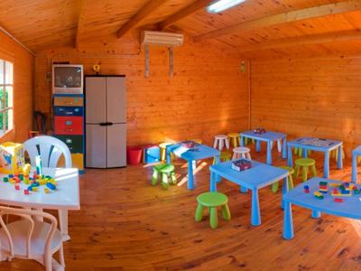 Sala giochi luminosa in legno con tavoli colorati, sedie e giocattoli vicino a grandi finestre.