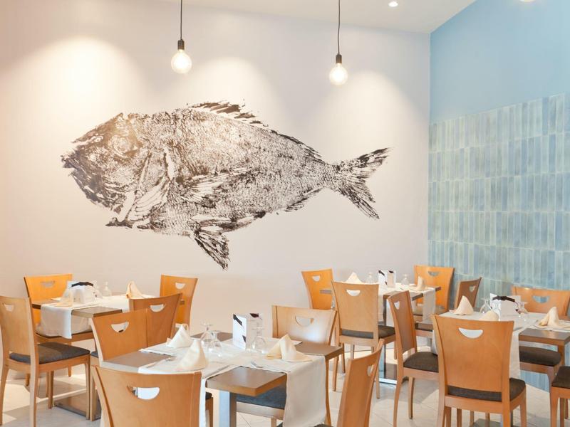 Accogliente area ristorante con sedie in legno, tavoli bianchi e parete decorata con pesce.