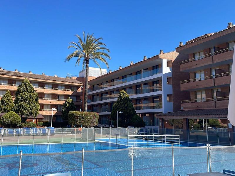 Cortile soleggiato dell'hotel con piscina, palma e balconi su più piani sotto un cielo azzurro chiaro.