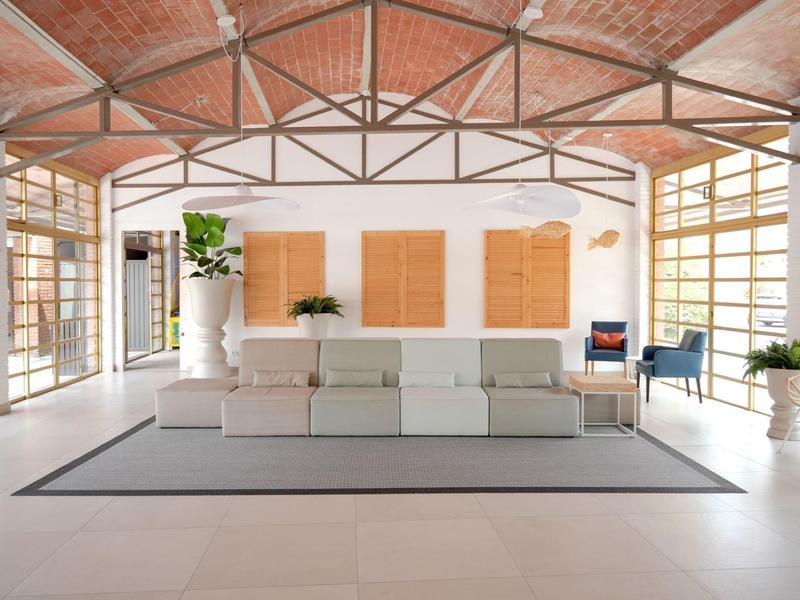 Ampio salotto con divani bianchi, piante, soffitto in legno e grandi finestre che lasciano entrare luce naturale.