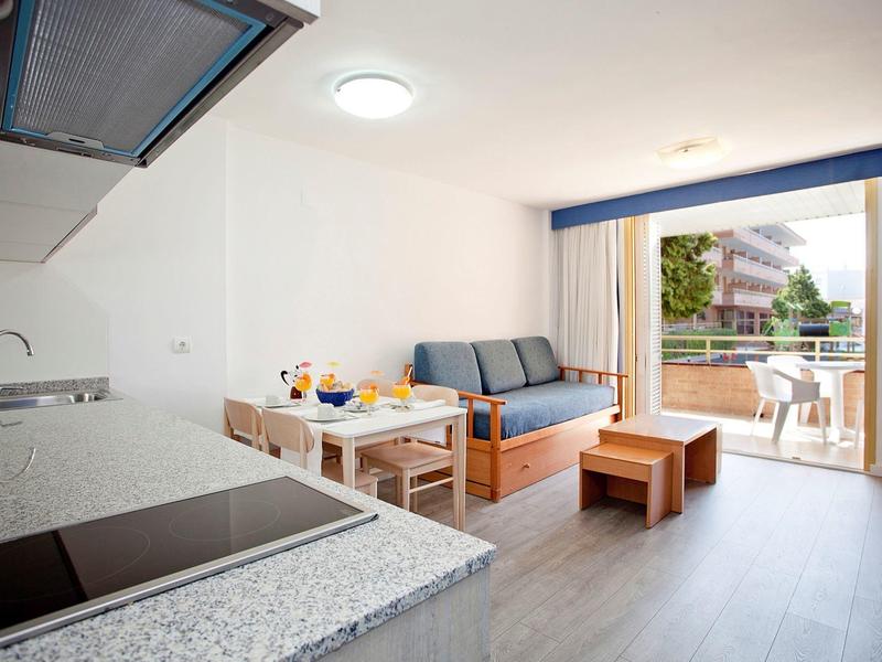 Camera d'hotel luminosa con angolo cottura, tavolo da pranzo, divano e balcone con vista sul verde.