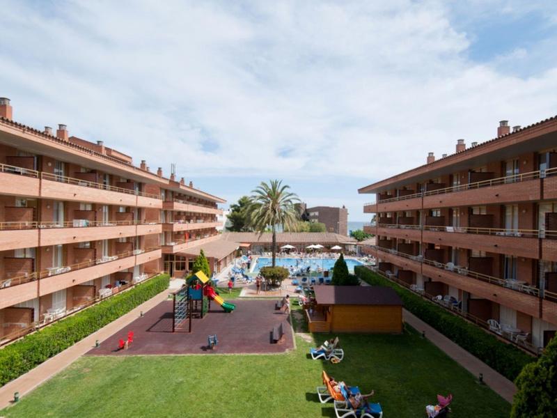 Resort con grande piscina, parco giochi, palme, lettini fra due lunghi edifici di appartamenti.