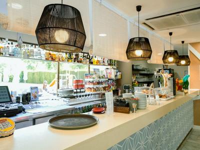 Bar d'hôtel moderne avec comptoirs blancs, suspensions noires et espace boissons bien approvisionné.