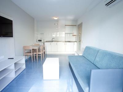 Salon lumineux et moderne avec kitchenette, canapé bleu, table à manger et téléviseur à écran plat.