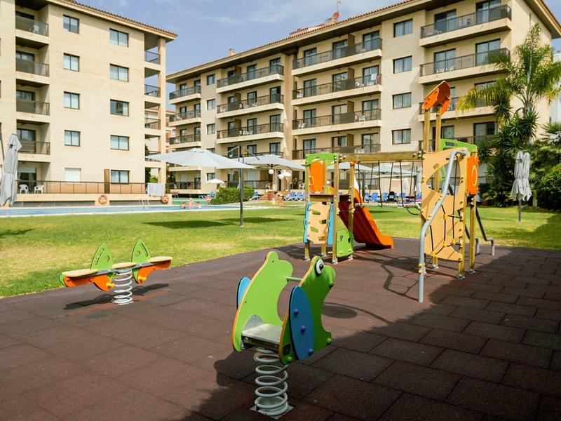 Aire de jeux avec toboggan et balançoire devant un immeuble d'hôtel à plusieurs étages sous le soleil.