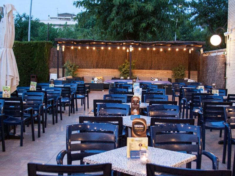 Restaurant en plein air avec des chaises noires, des tables et une zone éclairée à l'arrière-plan.