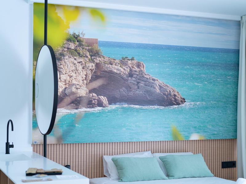 Pokój hotelowy z łóżkiem, zielonymi poduszkami i dużym zdjęciem klifu i morza na ścianie.